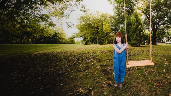 Child in blue overalls and brown hat standing next to a wooden swing in a park